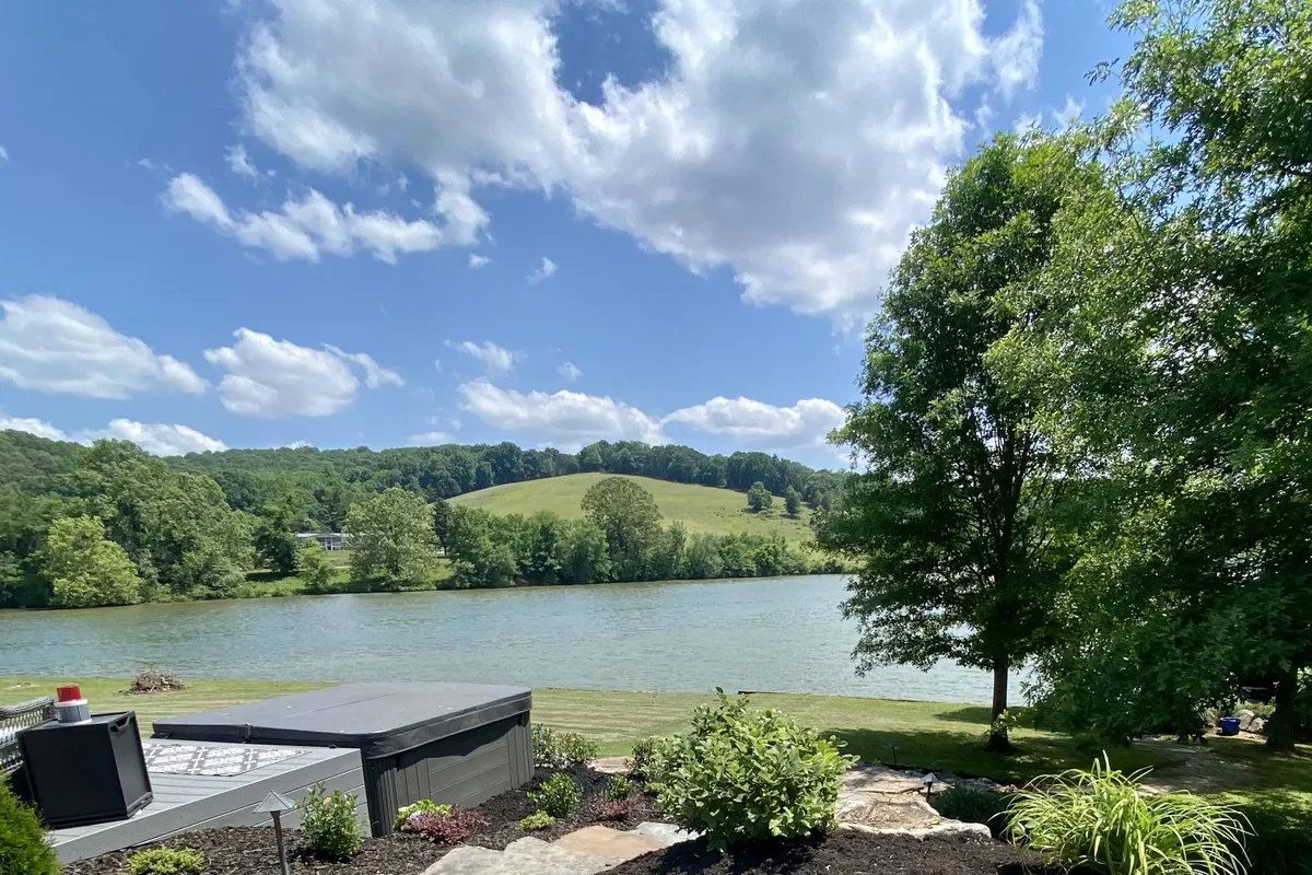 A peaceful lakeside view with green hills, surrounding trees, and a patio area set right by the water, inviting relaxation, picnics, or stargazing. The atmosphere is serene and private, fitting for either romantic moments or shared experiences with friends.