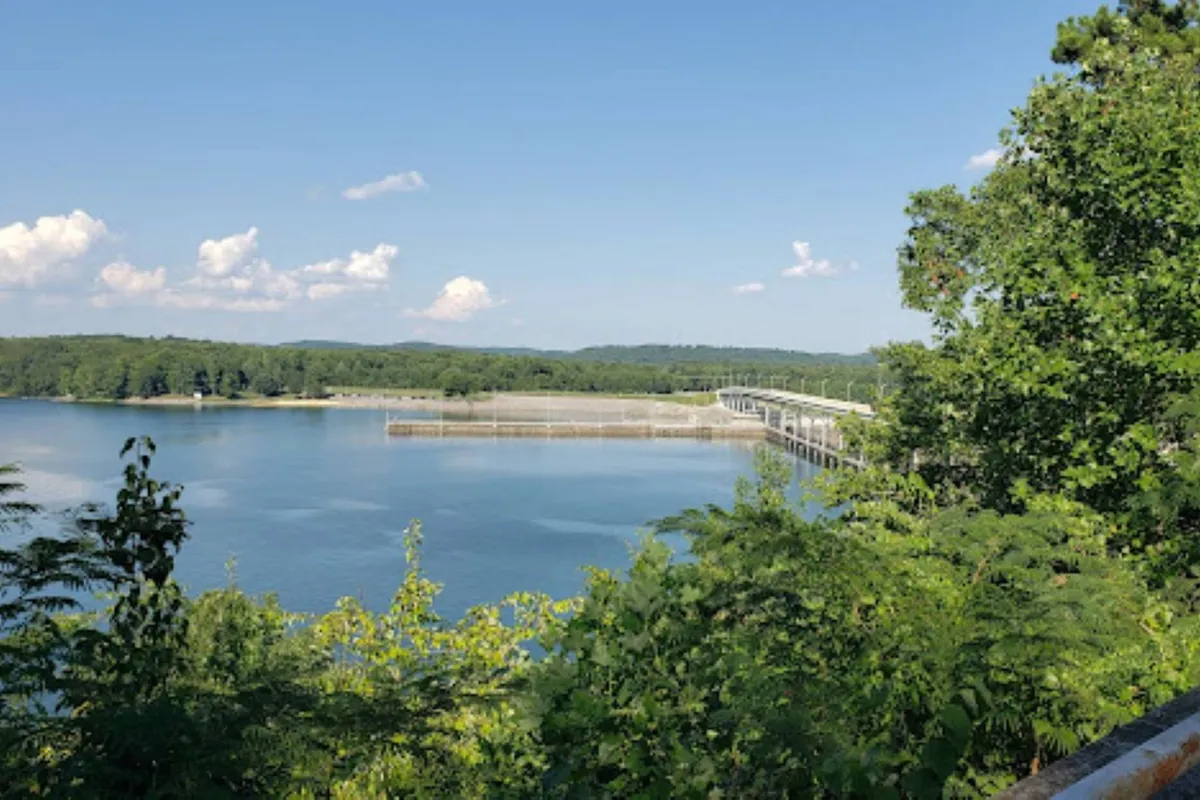 A peaceful lake scene featuring calm waters, a distant bridge, and abundant green foliage under a blue sky. The view evokes relaxation after a day of adventures, and suggests opportunities for fishing, boating, or simply unwinding at the water's edge.