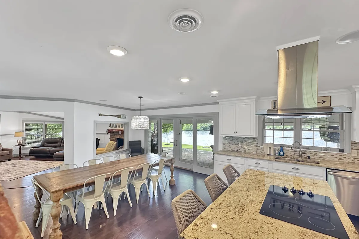A welcoming dining space anchored by a large table, with a kitchen island in the background and sliding glass doors that open to an outdoor deck. The room is designed for family meals and entertaining, with views that hint at the proximity to the lake.