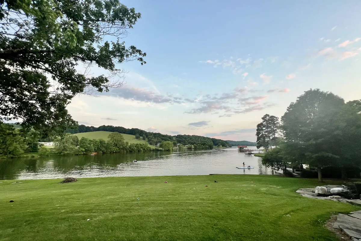 A calm lake stretches out in the foreground, bordered by a lush green hillside and distant trees. A kayaker glides peacefully across the water, embodying the sense of lakeside adventure and recreation. The scene hints at multiple possibilities for water activities such as kayaking, fishing, and enjoying the tranquil setting.