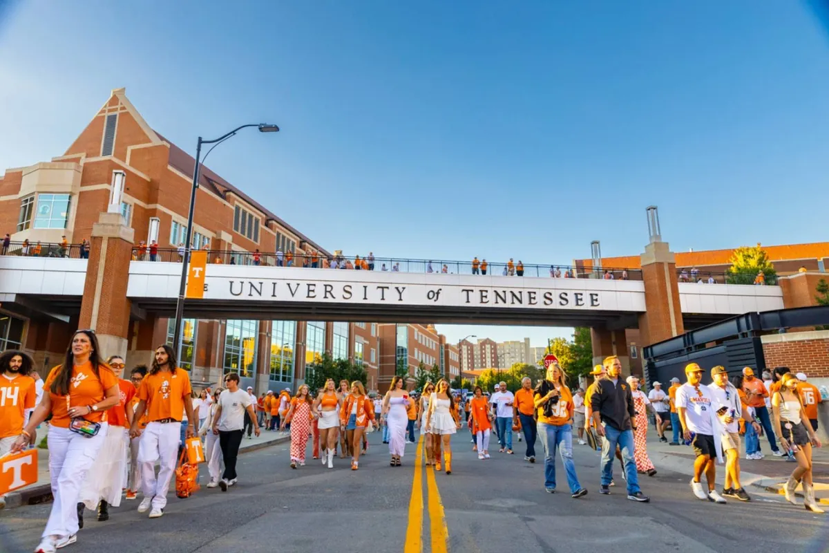 A vibrant scene outside the University of Tennessee, showcasing distinctive campus architecture, a lively crowd dressed in signature orange attire, and the university's prominent signage. The atmosphere is energetic and festive, capturing the essence of attending an iconic football game or campus event.