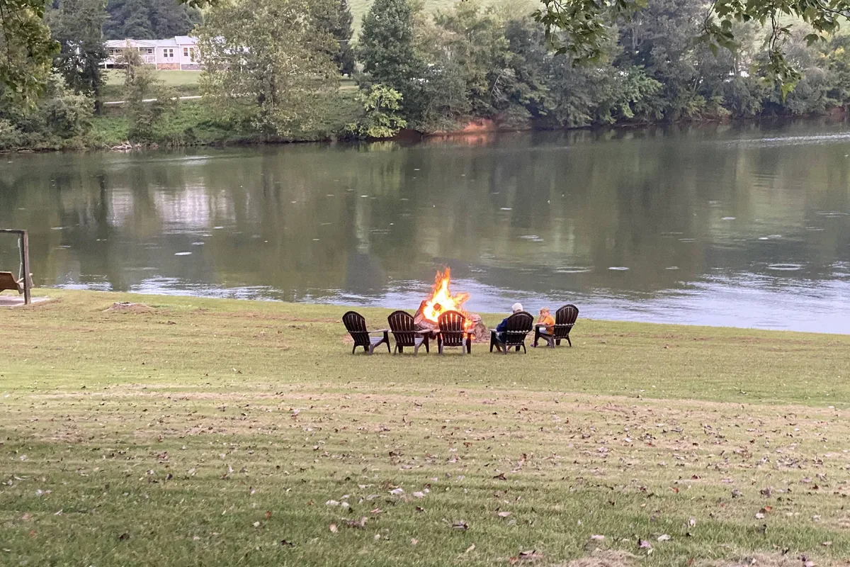 An inviting outdoor fire pit area with several lawn chairs arranged around a glowing fire, set against the tranquil backdrop of the lake. The open space hints at plenty of room for both intimate gatherings and group activities, with direct views out toward the water and the evening sky.