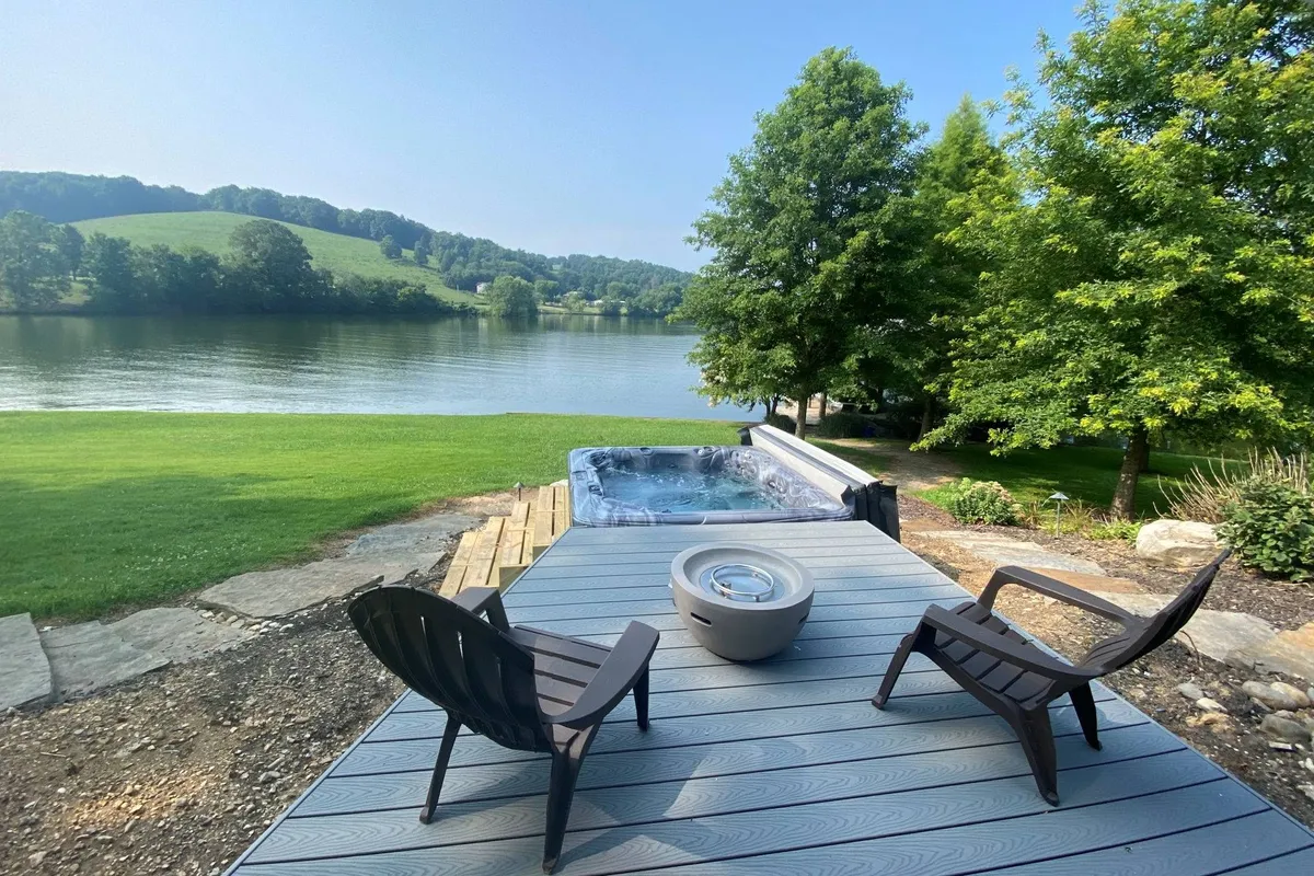 The hero image showcases a spacious lakeside backyard with a bubbling hot tub, inviting lawn chairs encircling a crackling fire pit, and sweeping views of tranquil Watts Bar Lake. The space is set up for relaxation, social gatherings, and outdoor enjoyment, all framed by serene natural surroundings that hint at breathtaking sunsets and peaceful stargazing.