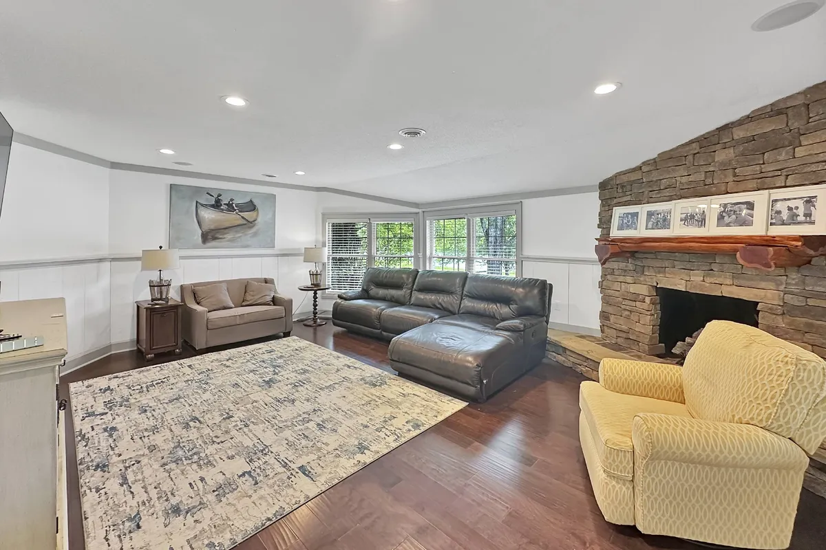 A spacious living room featuring a prominent stone fireplace as a warm focal point. The room is furnished with a large leather sectional sofa and an art piece above the fireplace, set on a cozy area rug. The space exudes comfort and is clearly designed for family gatherings, relaxation, or movie nights.
