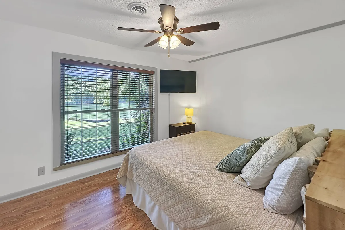 A tranquil bedroom scene featuring a king bed adorned with decorative pillows, a window with blinds allowing soft natural light, a nightstand with a lamp, and a TV mounted on the wall. The space feels airy and peaceful, ideal for rest and relaxation after a day outdoors.