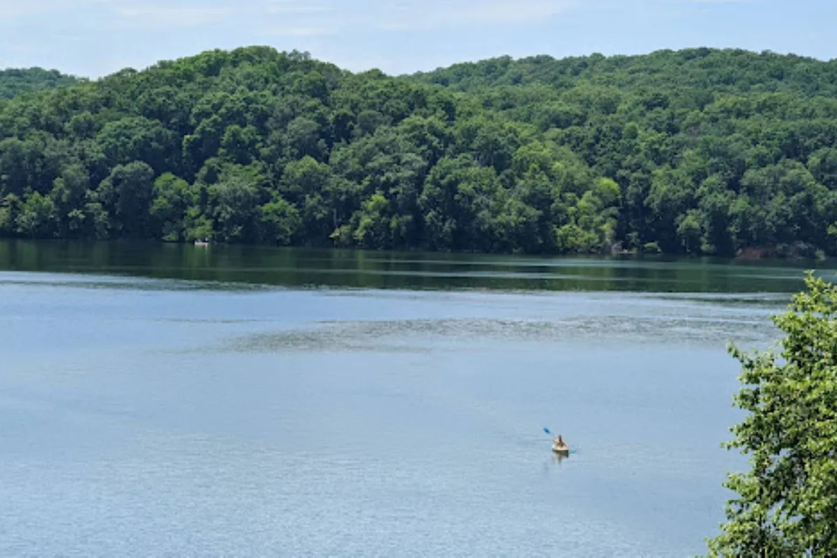 A calm lakeside scene featuring a lush green hillside, a kayaker on the water, and wide open skies. The image invokes a sense of tranquility and the allure of water-based activities, such as kayaking or simply relaxing by the water's edge.