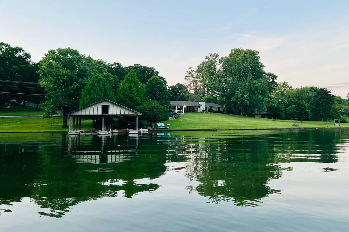 A wide shot shows the private house with direct frontage on the lake, a boathouse, and surrounding greenery. The tranquil water invites relaxation, while the property layout makes clear the immediate access to lakeside fun and amenities.